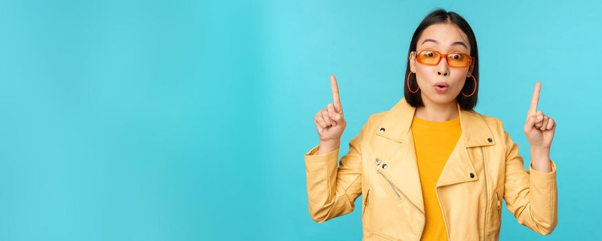 Enthusiastic asian girl in sunglasses, points fingers up, shows banner or logo on top, stands over blue background. Copy space
