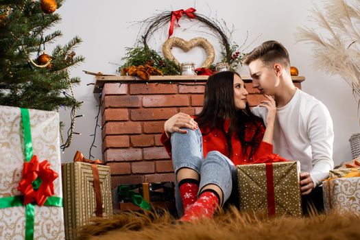 The bride and groom, sitting by the Christmas tree, communicate their feelings in body language. Lovers flirt in a decorated living room at home. December Christian holidays.