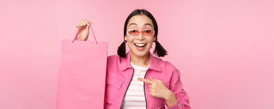 Shopping. Stylish asian girl in sunglasses, showing bag from shop and smiling, recommending sale promo in store, standing over pink background.