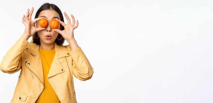 Funny asian girl holding tangerine on eyes and smiling, making playful grimaces, standing over white background. Copy space