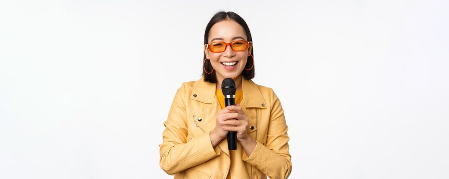 Stylish asian girl in sunglasses, singing songs with microphone, holding mic and dancing at karaoke, posing against white background.