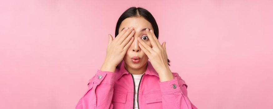 Close up portrait of young asian girl looking surprised, express amazement and wonder, peeking through fingers, standing over pink background.