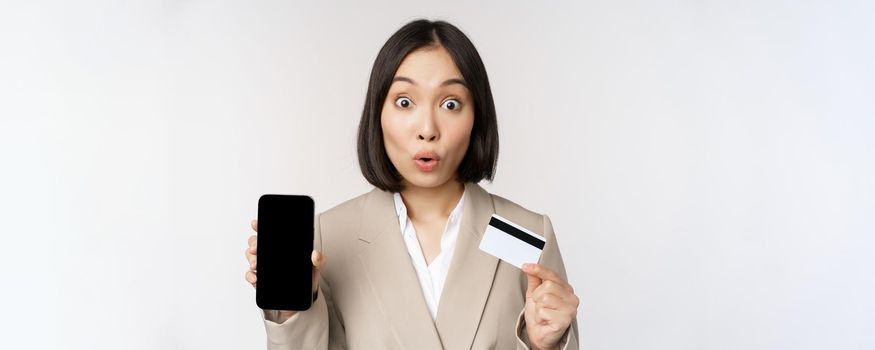 Corporate woman with happy, enthusiastic face, showing credit card and smartphone app screen, standing in suit over white background.