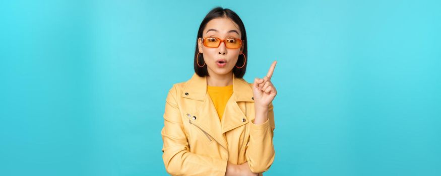 Excited asian girl raising finger, suggesting smth, eureka sign, has plan, standing in sunglasses over blue background.