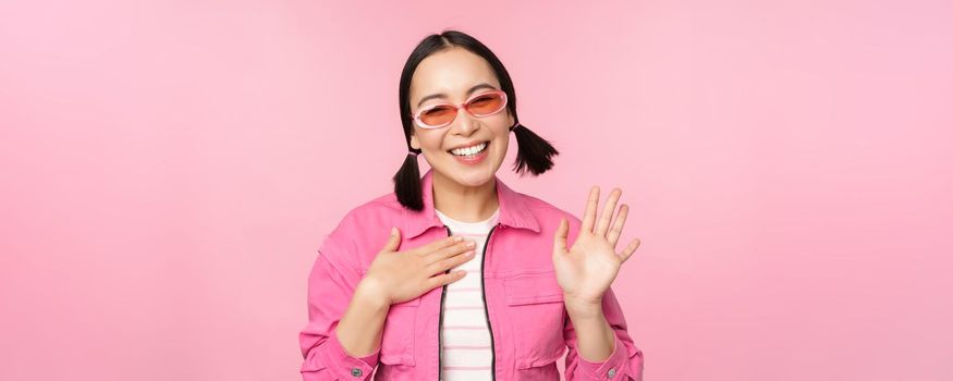 Portrait of friendly asian teen girl in sunglasses saying hello, waves her hand and smiles, greets you, hi gesture, stands over pink background.