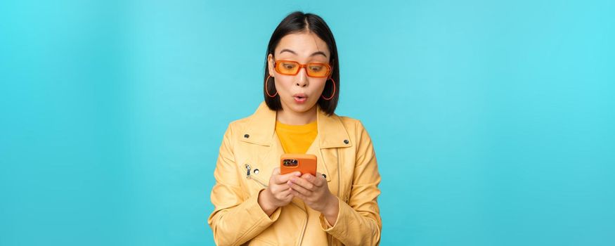 Portrait of asian girl in sunglasses, looking at smartphone screen with impressed, amazed face expression, wow face, standing over blue background.