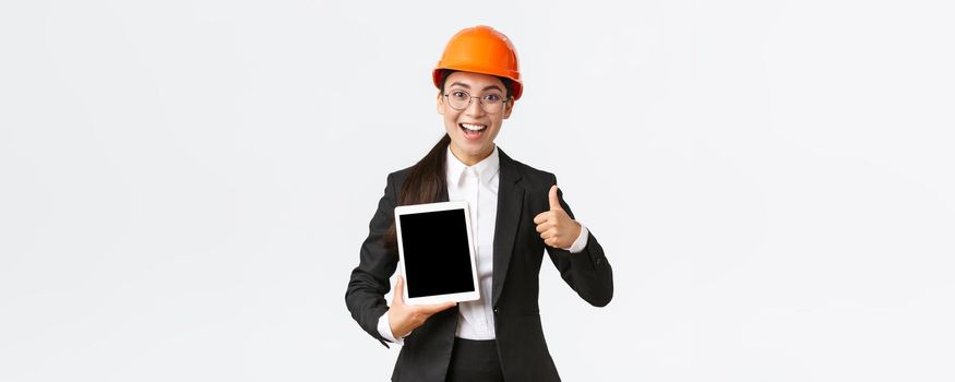 Impressed young female realtor, estate agent showing building during construction, wear safety helmet and business suit, holding digital tablet and make thumbs-up, recommend purchase.