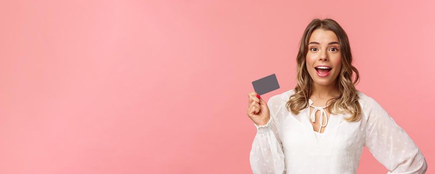 Close-up portrait of excited pretty young blond woman in white dress, holding credit card, showing people how she invests, look camera amused, smiling recommend shopping app, pink background.