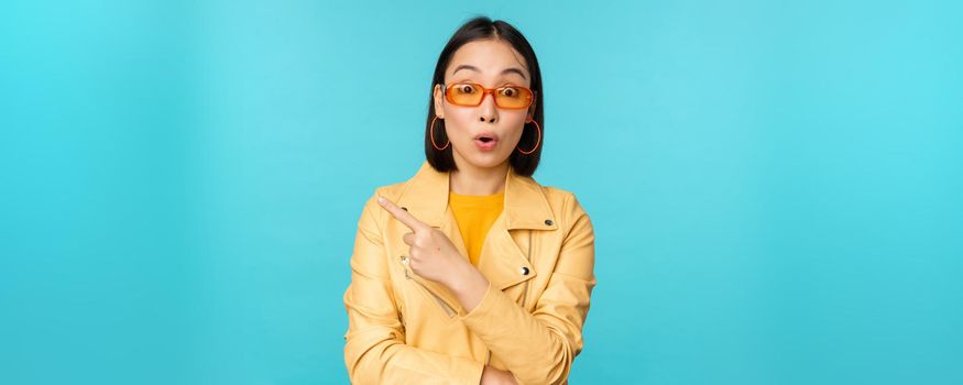 Image of asian woman looks intrigued, asks question about item or store, points finger left with surprised face expression, stands over blue background.
