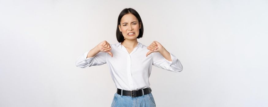 Dislike. Disgusted asian woman showing thumbs down and grimacing, commenting on smth bad, disagree, complaining, standing over white background.