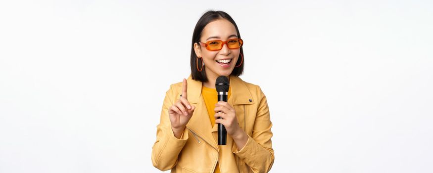 Portrait of beautiful asian woman in sunglasses, stylish girl singing, giving speech with microphone, holding mic and smiling, standing over white background. Copy space