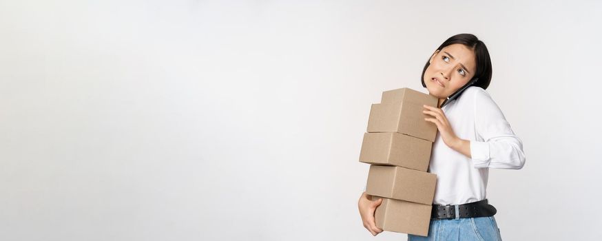 Young asian businesswoman answer phone call, talking on mobile while carrying pile of boxes with orders, standing over white background.