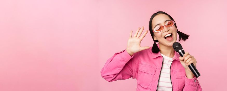 Happy beautiful asian girl singing with mic, using microphone, enjoying karaoke, posing against pink studio background.