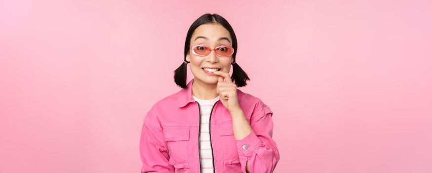 Stylish asian girl in sunglasses, looks thoughtful or intrigued, wants to try smth out, stands over pink background, pondering.