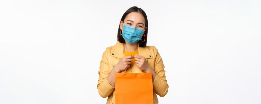Covid-19 and shop concept. Young asian stylish woman, wearing medical face mask, holding shopping bag, going in malls during pandemic, white background.