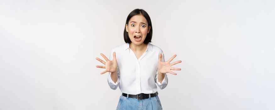 Asian woman looks at camera and screams in panic. Young korean girl looking anxious, panicking, shaking hands and shouting, standing over white background.