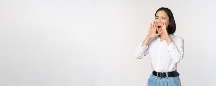 Image of young asian woman calling for someone, shouting loud and searching around, standing against white background.