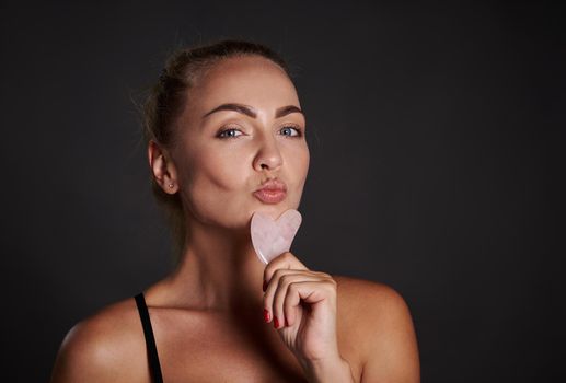 Beautiful Caucasian woman with healthy fresh smooth tanned skin posing with a gua-sha, ayurvedic jade stone massager for facial lifting and lymphatic drainage massage, isolated over black background