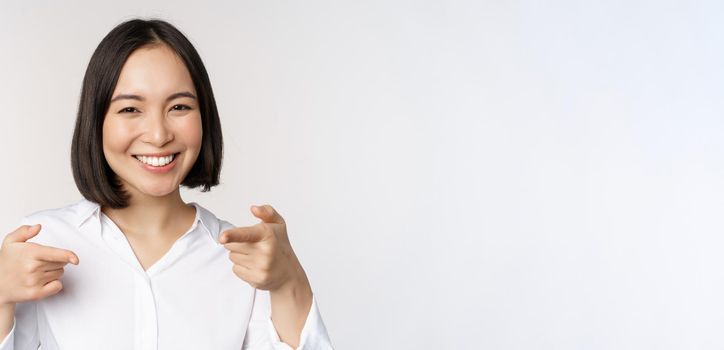 Close up of enthusiastic young woman smiling, pointing fingers at camera, choosing you, inviting and recruiting people, standing over white background.