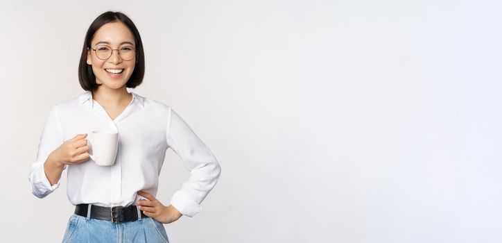 Happy young energetic asian woman smiling, drinking, holding cup mug of coffee, standing confident against white background.