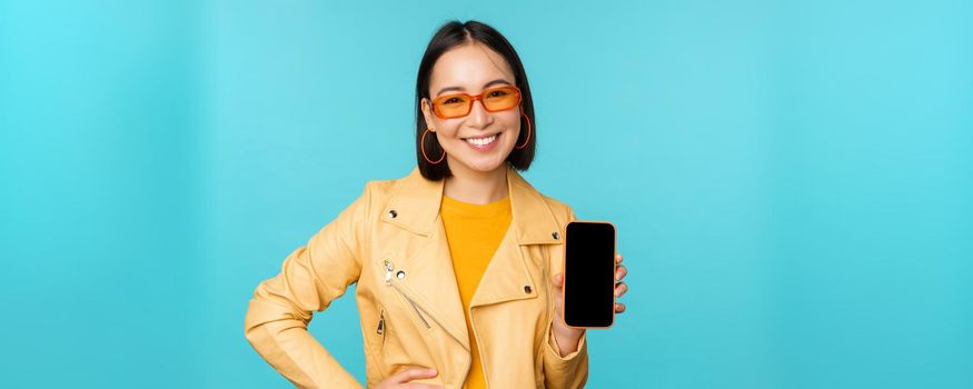 Image of young stylish asian female model in trendy sunglasses, showing mobile phone screen, application on smartphone interface, standing over blue background.