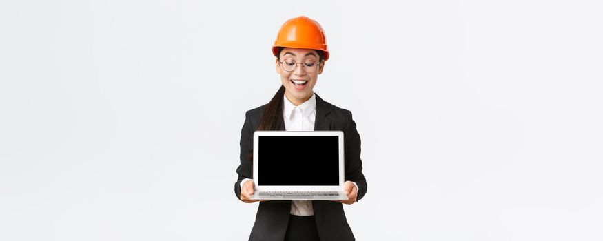 Impressed happy asian female chief engineer showing great results of enterprise to investors, standing in safety helmet and business suit, holding laptop and look pleased at computer screen.