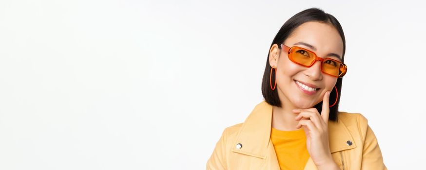 Close up portrait of asian woman thinking, wearing sunglasses and smiling, looking up thoughtful, standing over white studio background. Copy space