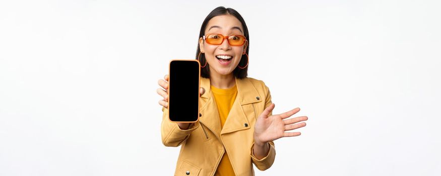 Enthusiastic asian female model, showing smartphone app interface, online store or website on mobile phone screen, standing over white background.