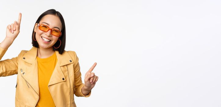Smiling asian brunette woman in sunglasses, pointing fingers sideways, left and right, showing variants, laughing and dancing, wearing sunglasses, white background.