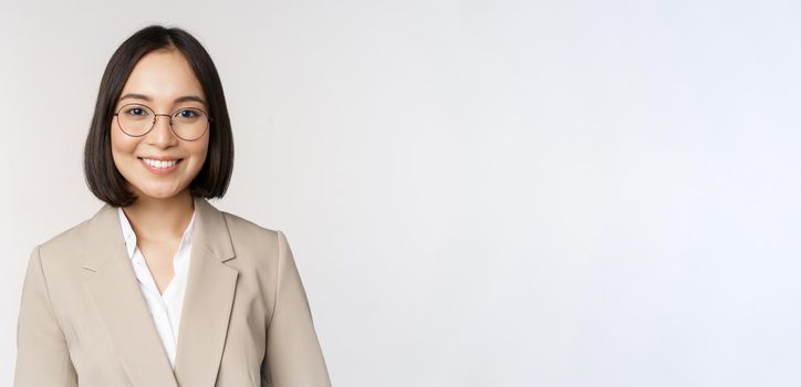 Portrait of young asian saleswoman in glasses, wearing beige suit, smiling and looking confident at camera, white background.