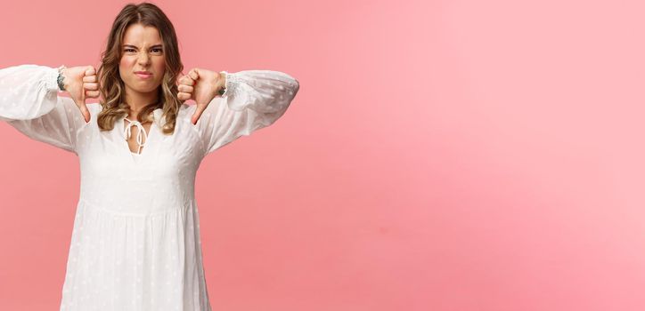 Portrait of picky young blond caucasian woman in white dress over pink background expressing dislike, show thumbs-down and grimacing in aversion, disappointed with bad quality.