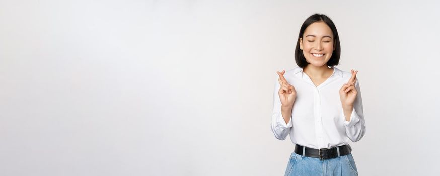 Portrait of young korean woman, asian girl cross fingers and praying, making wish, anticipating, waiting for results, standing over white background.