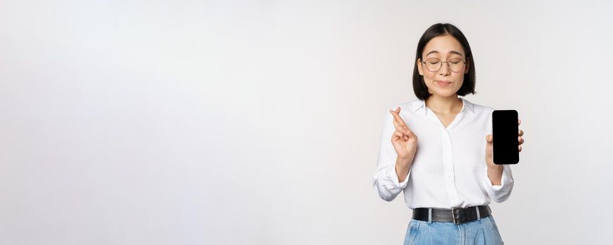 Hopeful young asian woman, showing mobile phone screen, app interface and fingers crossed, hoping for smth, making wish or paying, standing over white background.