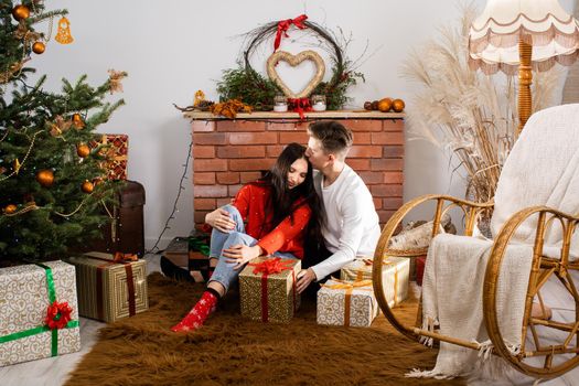 The bride and groom, sitting by the Christmas tree, communicate their feelings in body language. Lovers flirt in a decorated living room at home. December Christian holidays.