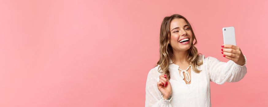 Close-up portrait of cheerful upbeat smiling blond girl, wearing white dress, laughing as record video, calling friend on mobile application, taking photo, selfie on smartphone, pink background.