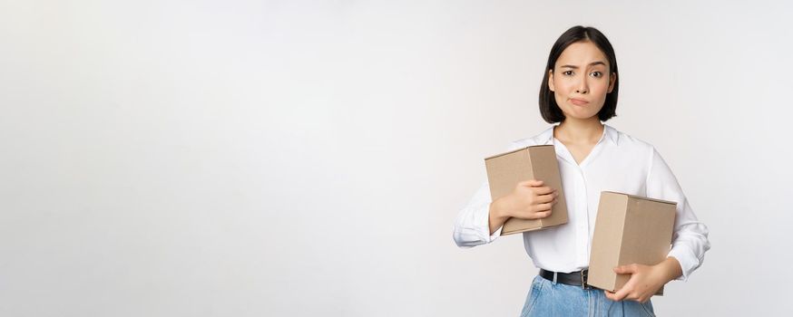 Complicated young asian woman holding two boxes, looking doubtful at camera, standing over white background puzzled.