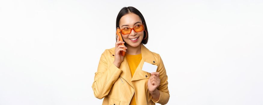 Stylish asian female model, talking on smartphone and showing credit card, standing over white background. Copy space