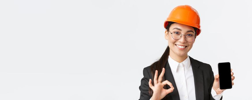 Satisfied young female asian engineer, architect in suit and safety helmet showing mobile phone screen and make okay gesture, approve, recommend application, white background.