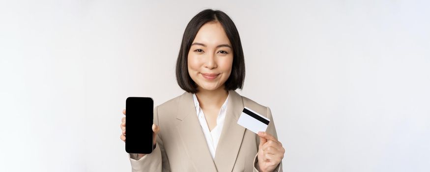 Smiling corporate woman in suit, showing mobile phone screen and app on mobile phone, smartphone screen, standing over white background.