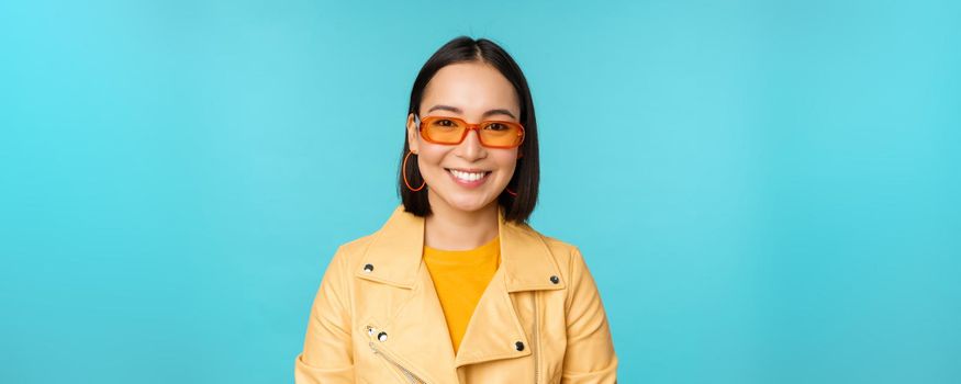 Close up portrait of smiling stylish asian woman in sunglasses, looking happy at camera, posing in trendy yellow jacket, blue background.