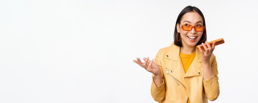 Image of happy asian girl talking on speakerphone, recording, translating her voice with mobile phone app, talking in smartphone dynamic, standing over white background.