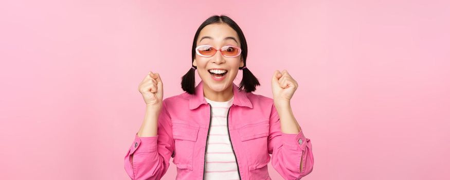 Portrait of excited japanese girl in sunglasses, celebrating, achieve goal, gasping amazed and smiling, standing over pink background.