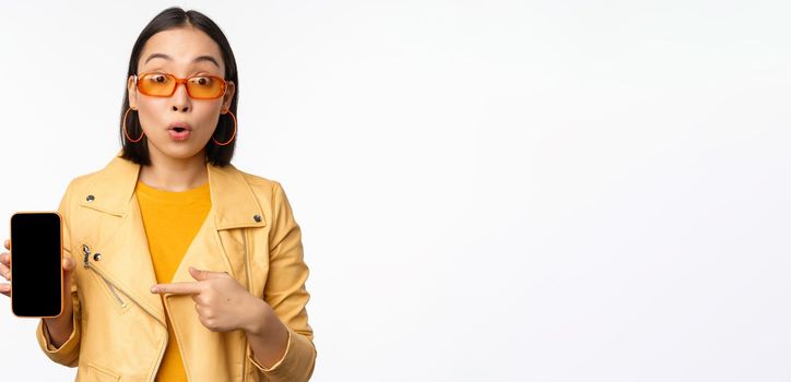 Portrait of stylish korean girl in sunglasses, smiling, pointing finger at smartphone screen, showing mobile phone application, standing over white background.