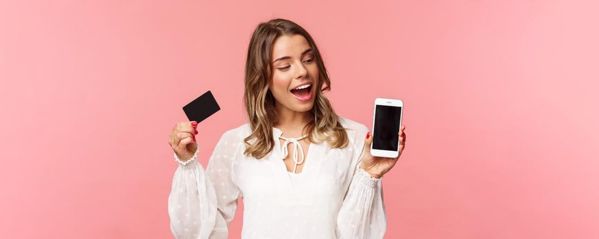 Finance, shopping and technology concept. Close-up portrait of excited good-looking blond girl in white dress, renew her spring wardrobe with buying clothes online via smartphone, hold credit card.