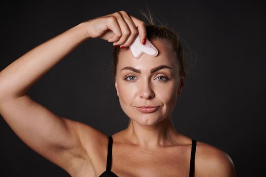 Studio shot. Beauty portrait of an attractive Caucasian woman massaging her face with a gua-sha jade stone massager. Anti-aging, anti-wrinkles, rejuvenating and anti-puffiness concept