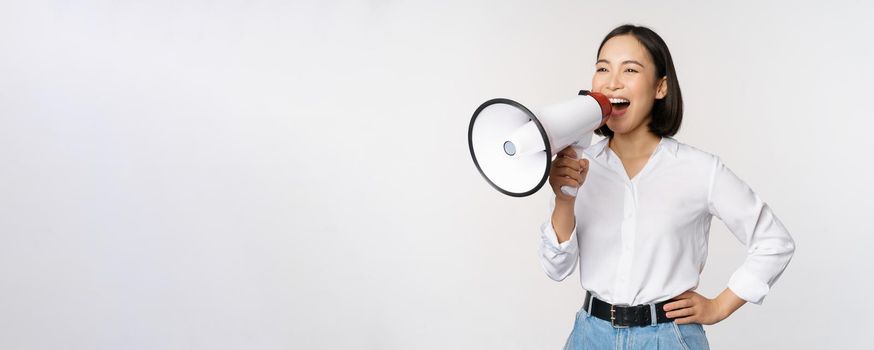 Image of young woman, korean activist, recruiter screaming in megaphone, searching, shouting at loudspeaker, standing over white background.