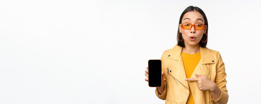 Portrait of stylish korean girl in sunglasses, smiling, pointing finger at smartphone screen, showing mobile phone application, standing over white background.