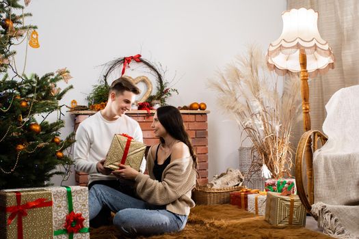 Two lovers are sitting by the Christmas tree on Christmas. Decorated Christmas tree and fireplace in the living room at home.