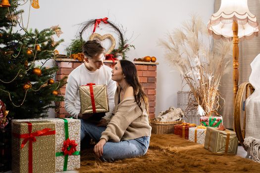 Two lovers are sitting by the Christmas tree on Christmas. Decorated Christmas tree and fireplace in the living room at home.