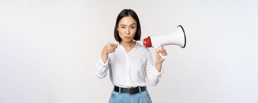 Image of modern asian woman with megaphone, pointing at you camera, making announcement, white background.